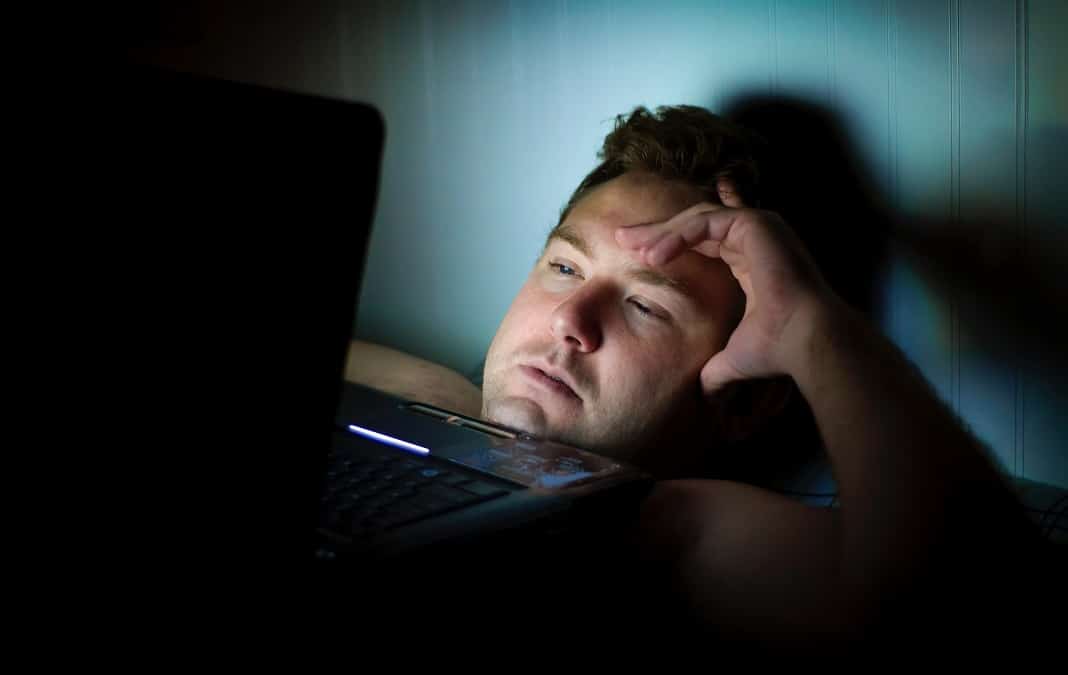 Tired man using a laptop while laying in bed.