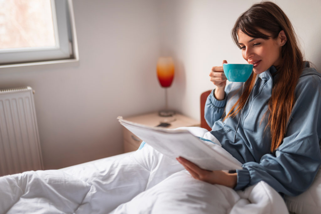 A young woman drinking coffee and reading the newspaper in bed.