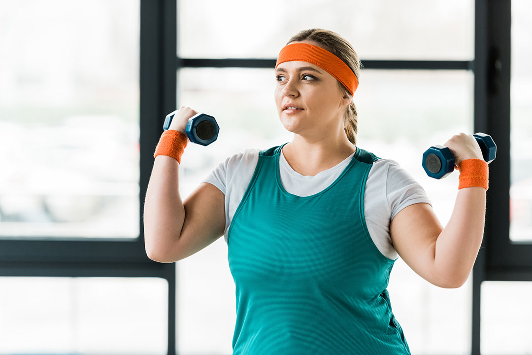 Plus size woman in blue tank top exercising in gym with dumbbells to lose weight