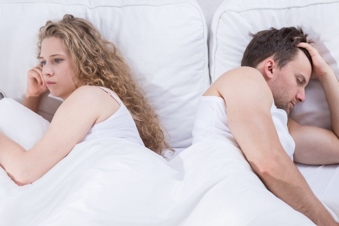 Possible couple candidate for a sleep divorce lay in bed with their backs to each other while the man sleeps and his female partner lays wide awake looking unhappy.