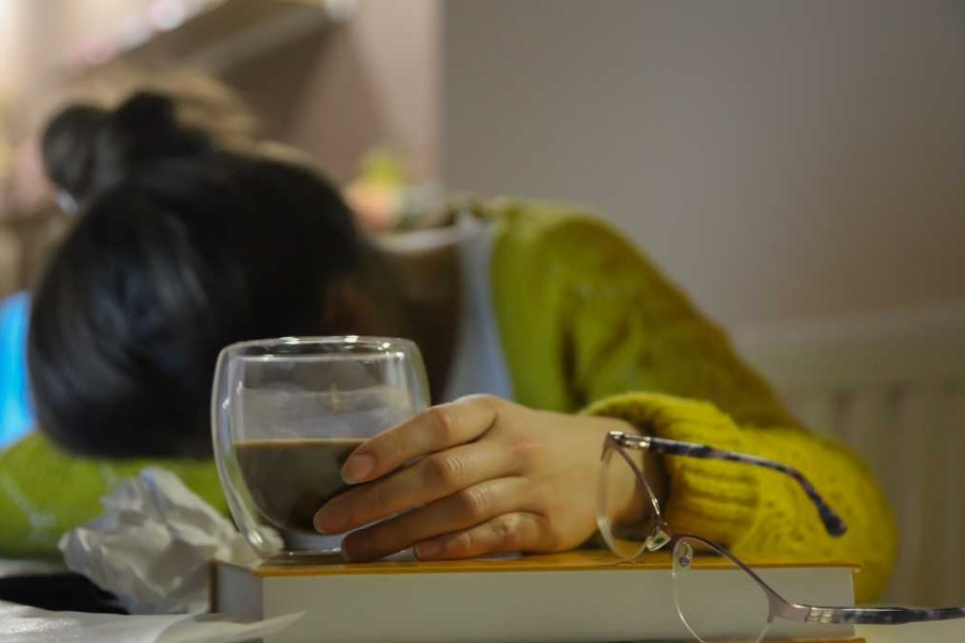 An exhausted female millennial has fallen asleep at her computer with her head resting on one arm while holding an alcoholic beverage with her other hand.