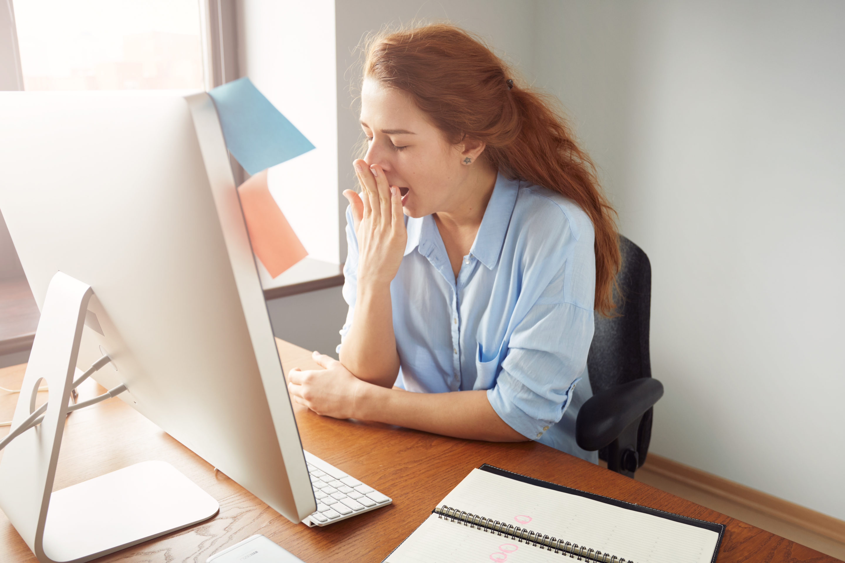 Tired woman working at her computer yawns in the afternoon during an afternoon slump.