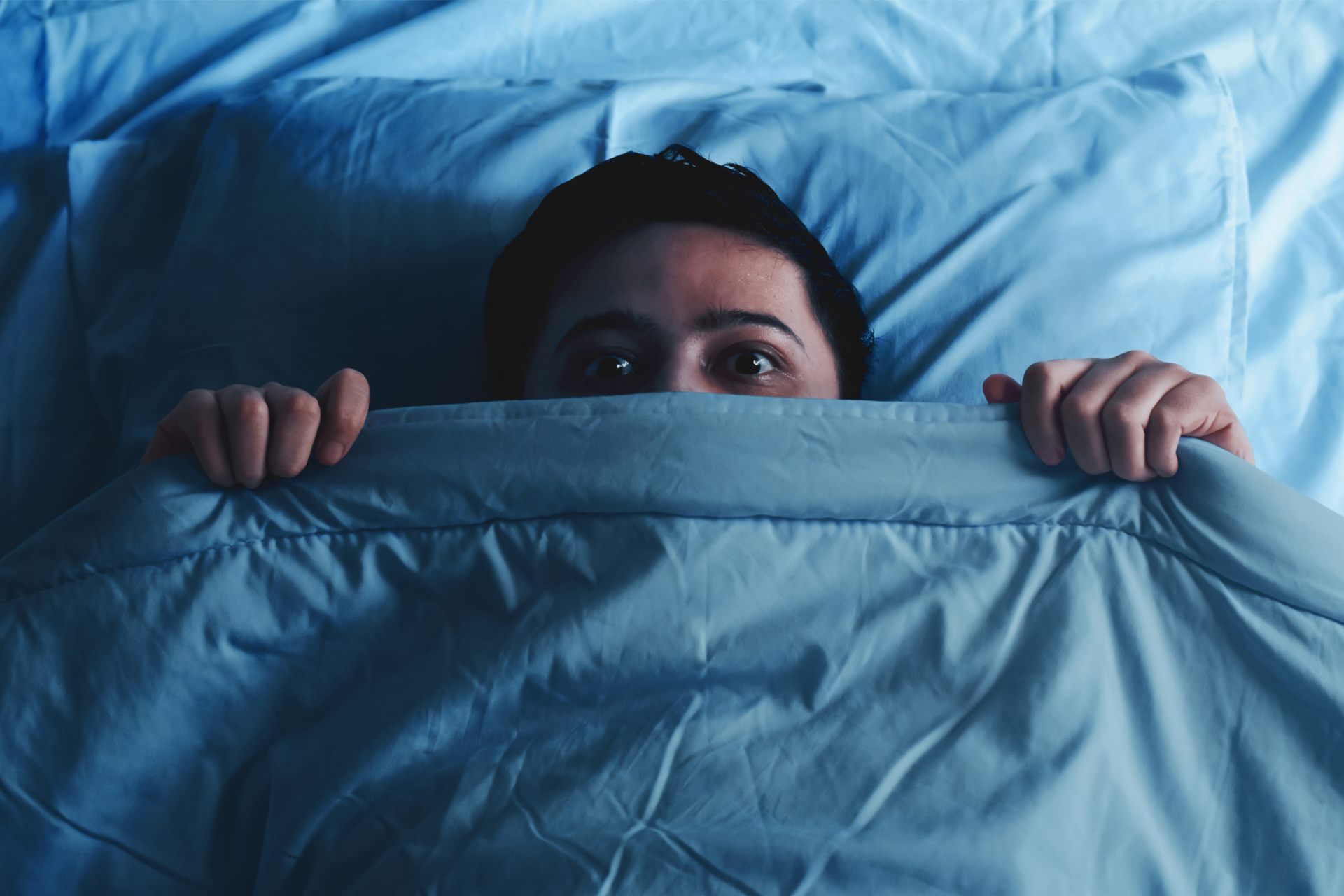 An anxious man laying in bed hiding his face under the sheets.
