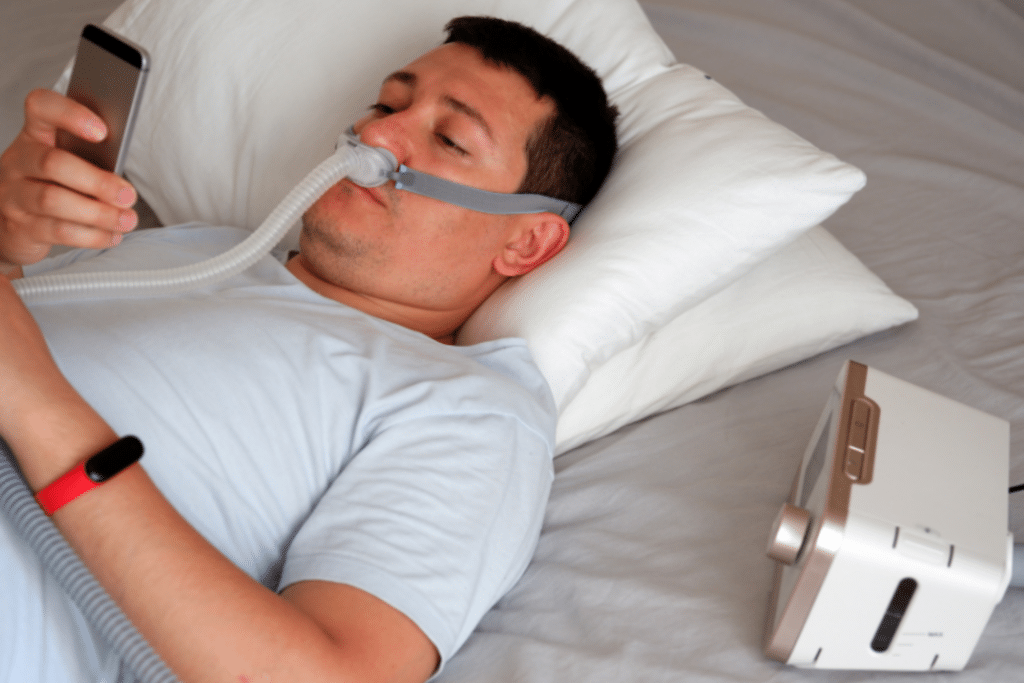 A man wearing a CPAP mask looking at his phone in bed.